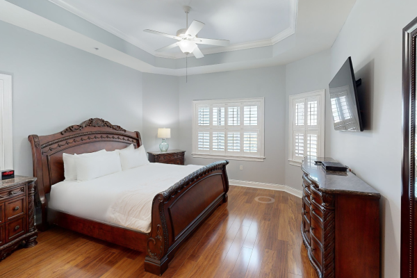 A bedroom with a large carved wooden bed, nightstands, lamps, hardwood floor, ceiling fan, windows, and a TV on the wall.