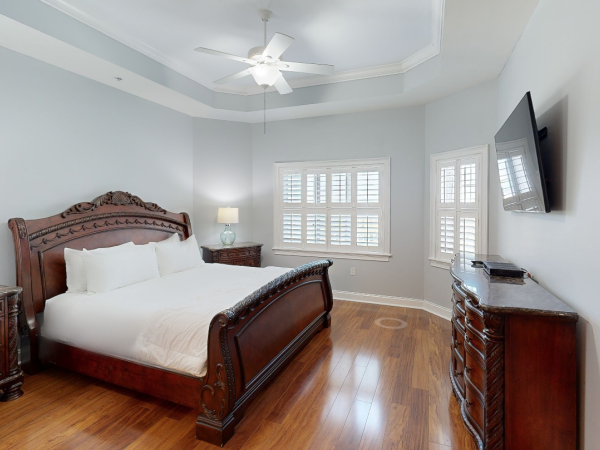 A bedroom with a large carved wooden bed, nightstands, lamps, hardwood floor, ceiling fan, windows, and a TV on the wall.