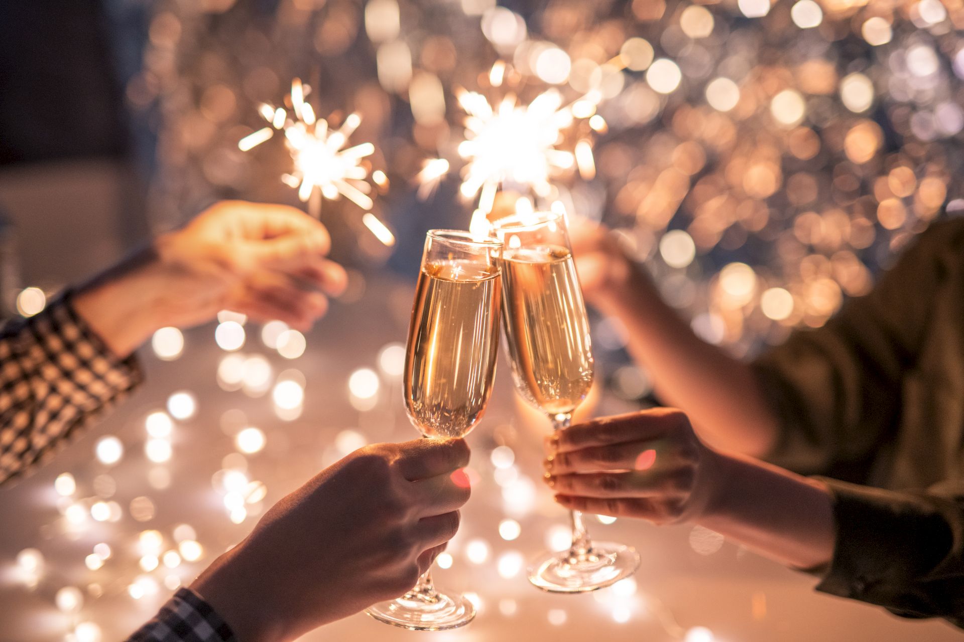 People are clinking champagne glasses and holding sparklers, celebrating in a festive atmosphere with sparkling lights in the background.