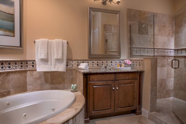A stylish bathroom features a large bathtub, a wooden vanity with a mirror, and a walk-in shower with beige tiles and elegant decor.