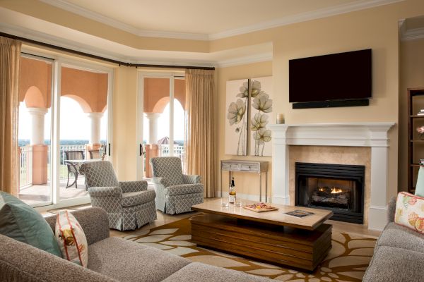 A cozy living room with a fireplace, wall-mounted TV, elegant armchairs, a coffee table, art, and a scenic balcony view.