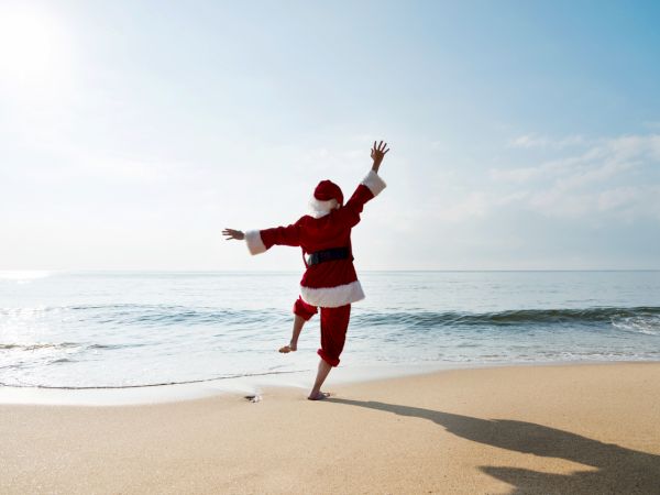 A person in a red Santa outfit joyfully runs and jumps on a sunny beach by the ocean.
