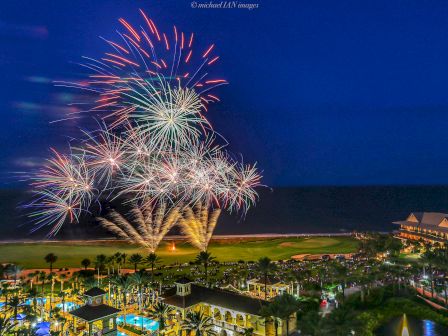 Fireworks light up the night sky above a resort with a pool, surrounded by palm trees and a building to the right.
