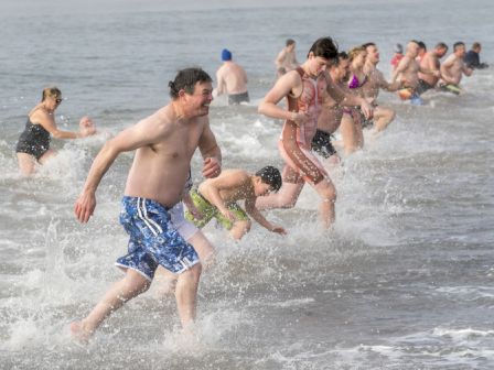 A crowd of swimmers runs and splashes in the ocean waves, laughing during a busy beach water game.