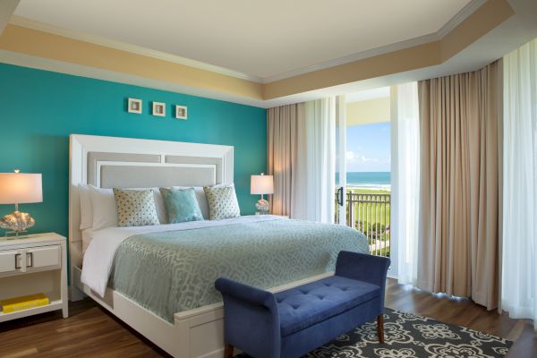 A cozy bedroom with a king-sized bed, turquoise accent wall, ocean view, and modern decor. Blue bench at the foot of the bed.