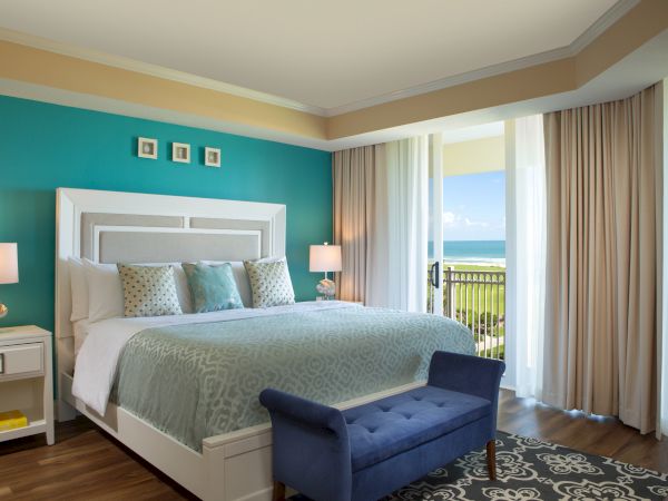A cozy bedroom with a king-sized bed, turquoise accent wall, ocean view, and modern decor. Blue bench at the foot of the bed.