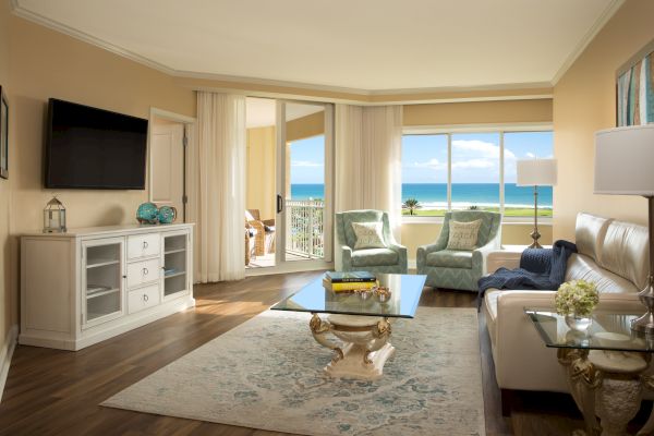 A modern living room with ocean view, featuring comfy chairs, a sofa, a TV, coffee table, and elegant decor against a serene backdrop.