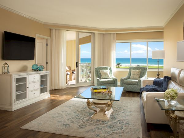 A modern living room with ocean view, featuring comfy chairs, a sofa, a TV, coffee table, and elegant decor against a serene backdrop.