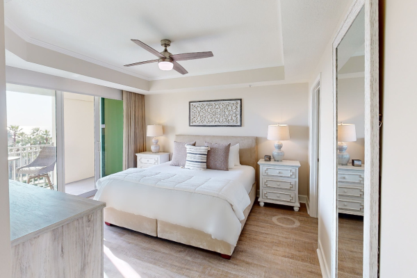 A cozy bedroom with a king-sized bed, two nightstands, a ceiling fan, and a balcony with outdoor chairs and a scenic view.