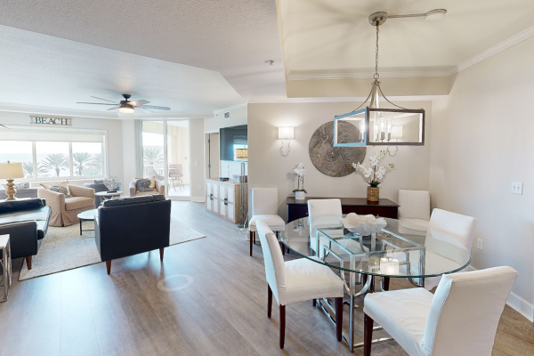 A contemporary living and dining area with a glass table, white chairs, modern chandelier, and a view of palm trees through large windows.