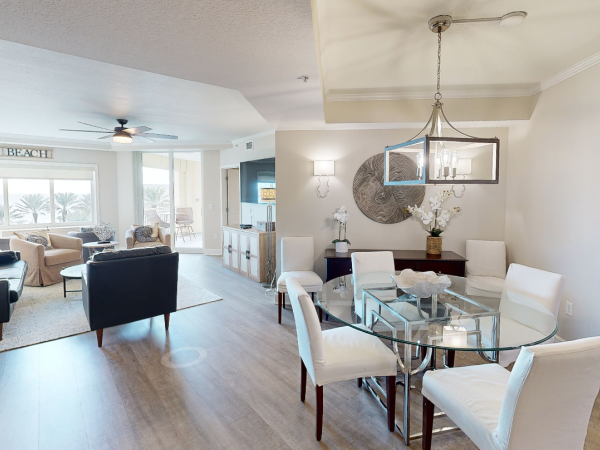 A contemporary living and dining area with a glass table, white chairs, modern chandelier, and a view of palm trees through large windows.