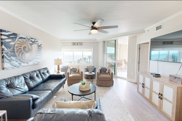 A cozy living room with sofas, armchairs, a large painting, a TV, and sliding doors leading to a balcony with a beach view.