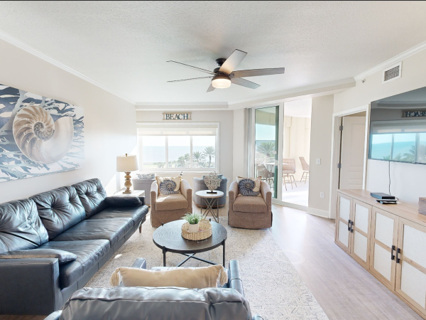 A cozy living room with sofas, armchairs, a large painting, a TV, and sliding doors leading to a balcony with a beach view.