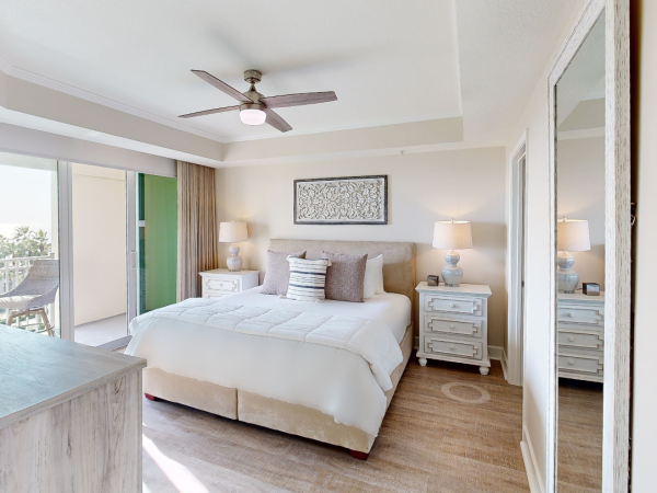 A cozy bedroom with a large bed, two bedside tables with lamps, a ceiling fan, and a balcony with a view.