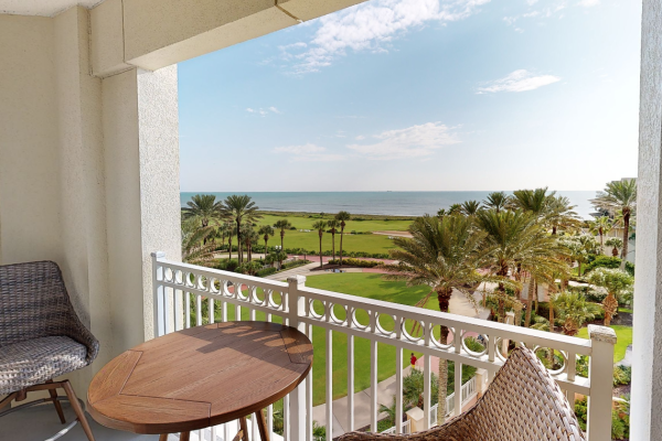 A balcony with a wicker chair and wooden table overlooks a lush garden with palm trees and a view of the ocean beyond.