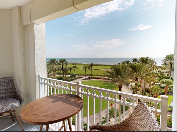 A balcony with a wicker chair and wooden table overlooks a lush garden with palm trees and a view of the ocean beyond.