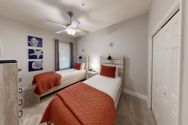 The image shows a bedroom with two single beds, red bedding, wall art, a ceiling fan, and a window between the beds.