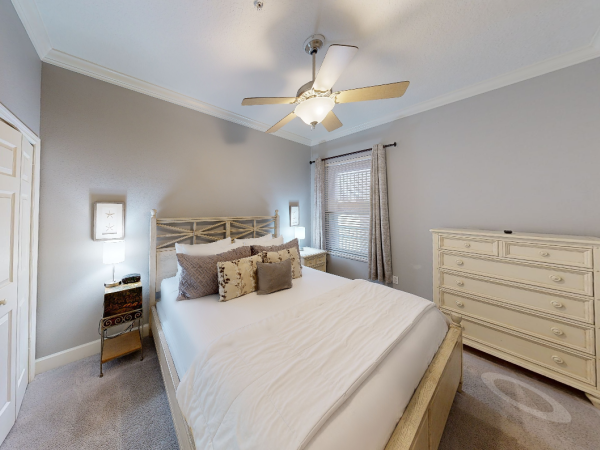 A cozy bedroom with a large bed, decorative pillows, two side tables with lamps, a ceiling fan, and a wooden dresser near the window.
