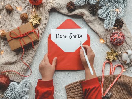 A child writes "Dear Santa" on paper in a red envelope, surrounded by festive decorations, candy canes, and a gift.