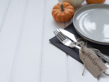 A Thanksgiving-themed table setting with a plate, utensils, small pumpkins, and a tag reading "Thanksgiving" on a white surface.