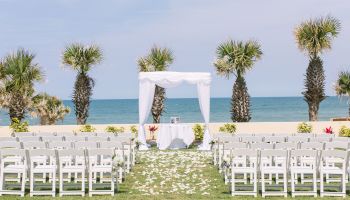 An outdoor wedding setup on a beach with white chairs, an altar, and palm trees in the background.