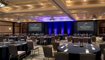 The image shows a conference room with round tables, chairs, stage, podium, and presentation screens ready for an event.