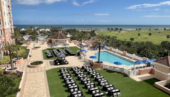 A resort area with a pool, sun loungers, and palm trees. The ocean is visible in the background under a clear blue sky.
