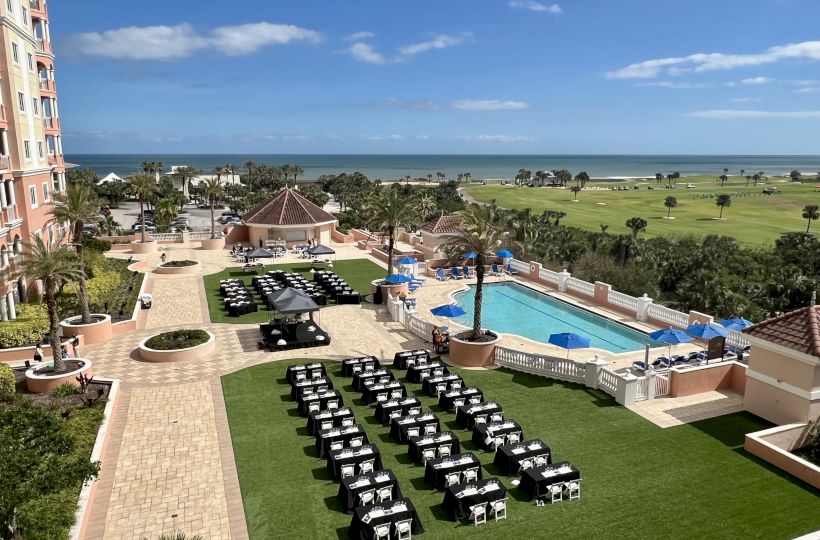 The image shows a resort with a pool, arranged seating on a lawn, palm trees, and a view of the sea under a clear blue sky.