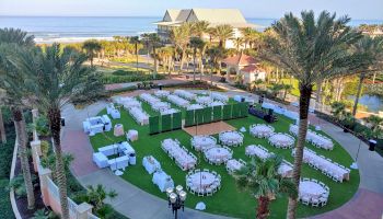 An outdoor event setup with tables and chairs is arranged on a lawn near the beach, surrounded by palm trees and a building in the background.