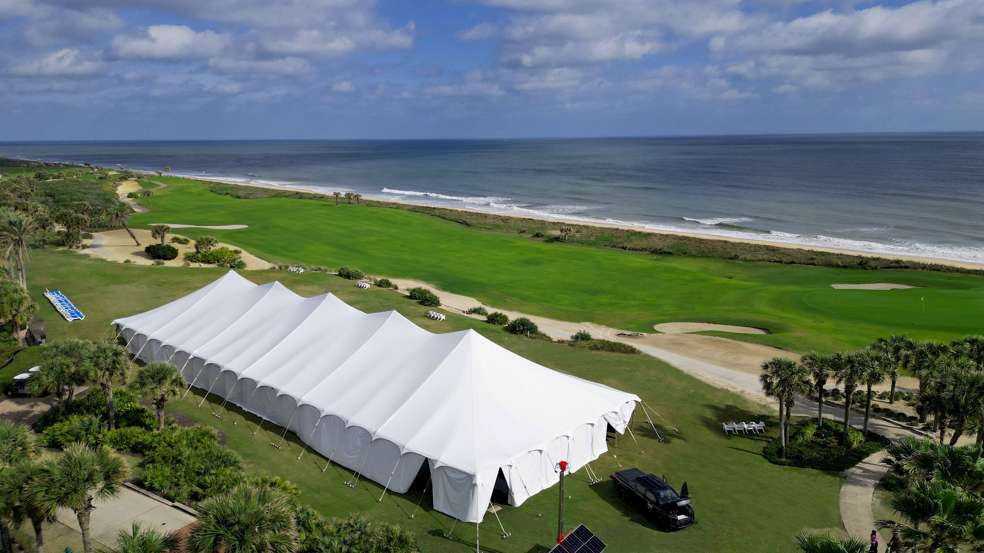A large white tent is set up on a grassy area near a beach, with the ocean in the background under a partly cloudy sky.