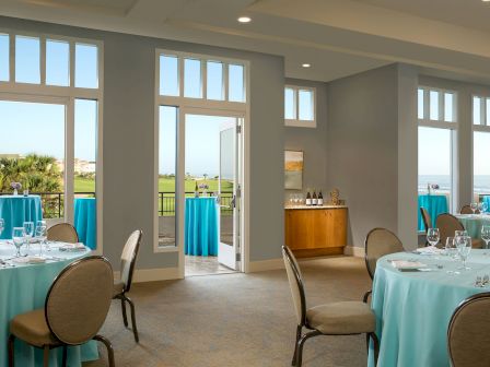 The image shows a bright event room with round tables covered in blue tablecloths set for dining, and an open door leading outside.