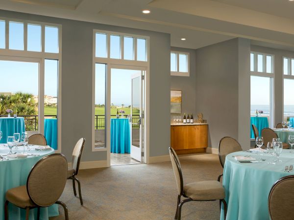 The image shows a bright event room with round tables covered in blue tablecloths set for dining, and an open door leading outside.
