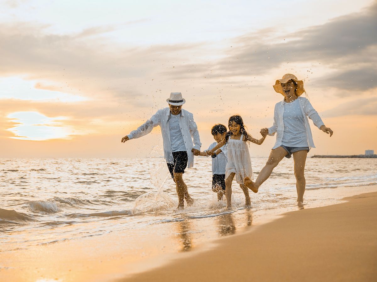 A family is joyfully walking along a beach during sunset, with two adults and two children holding hands and splashing in the water.