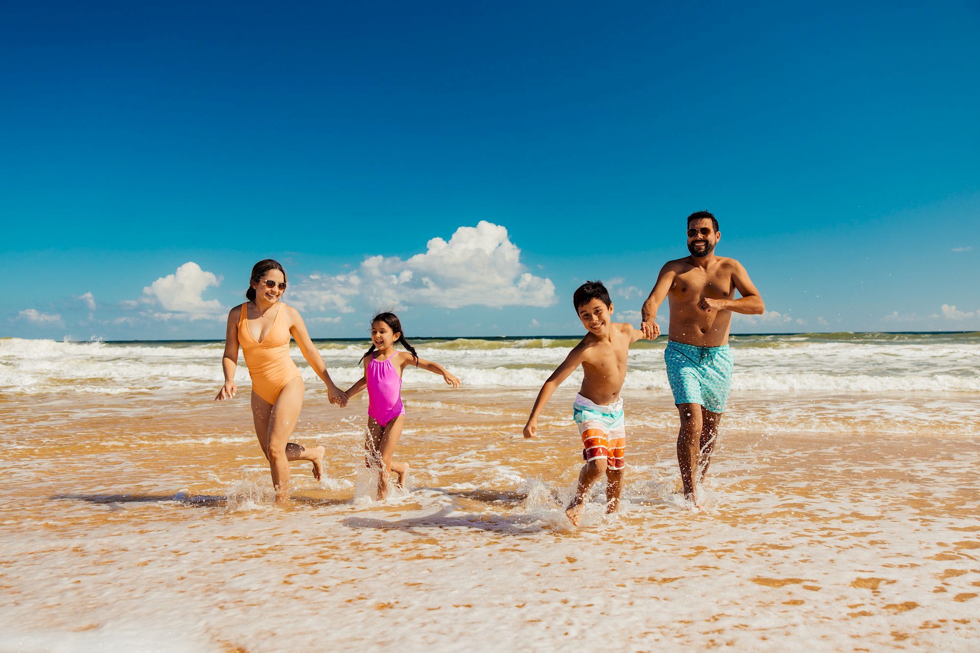 A family of four joyfully runs through shallow waves on a sunny beach, holding hands under a clear blue sky.