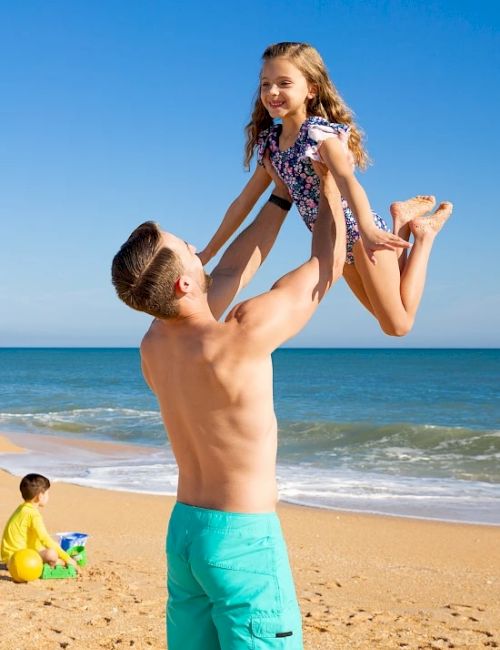 A man lifts a smiling girl in a swimsuit on a sandy beach. A child in yellow plays with a bucket near the water's edge.