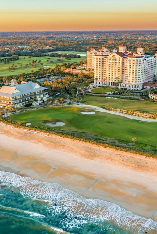 The image shows a beachfront with tall buildings, a golf course, and waves gently reaching the sand, under a clear sky.