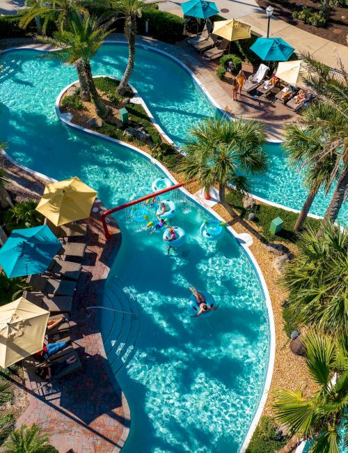 An aerial view of a winding pool surrounded by palm trees and lounge areas with umbrellas, creating a relaxing tropical atmosphere.