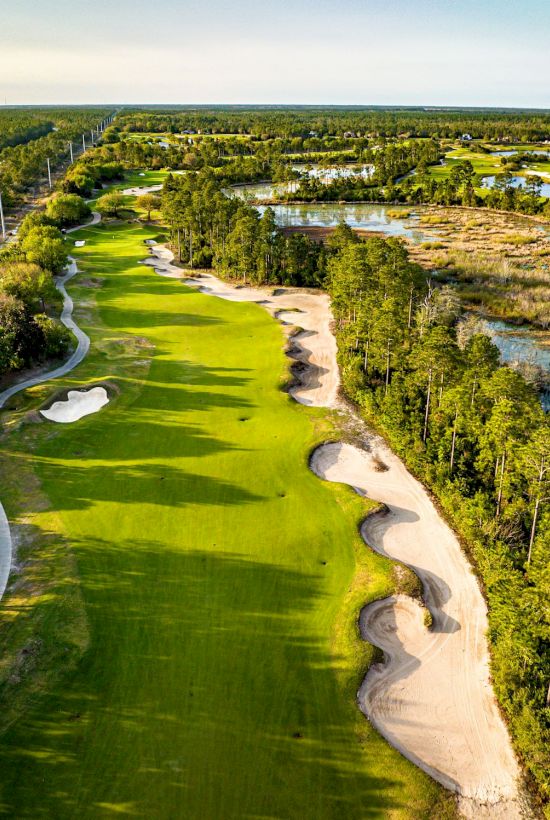 Aerial view of a golf course with lush greens, sand bunkers, and surrounding trees by a water body under a clear sky.