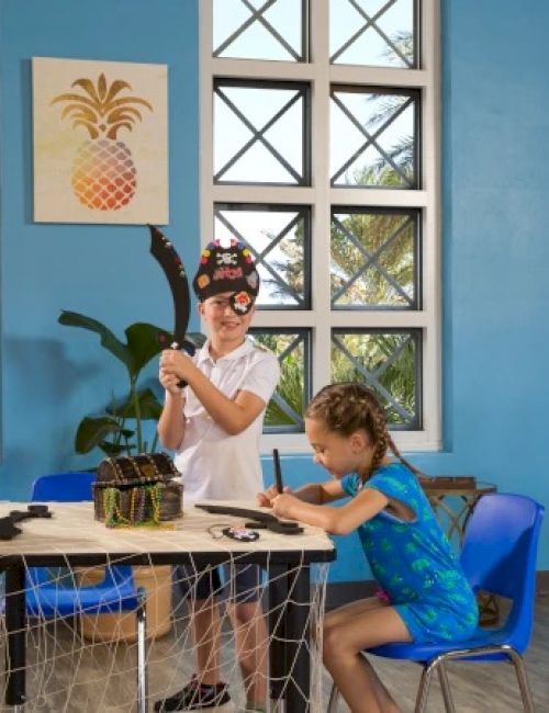 Children are playing in a classroom with a pirate theme, using props like a telescope and pirate hat, and drawing at a table.