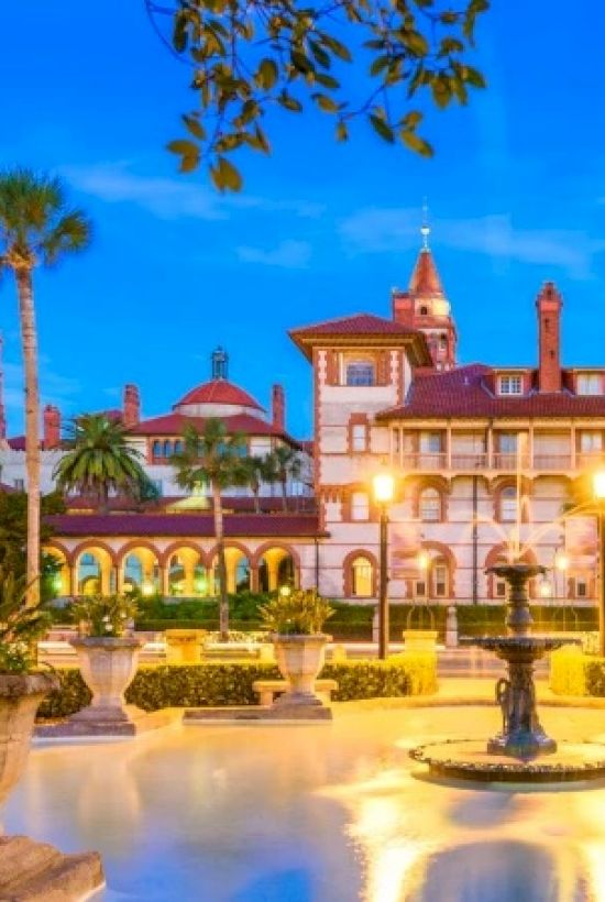 Historic buildings with palm trees and a fountain in the foreground at sunset.