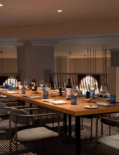 The image shows a set dining area with a long table, chairs, wine bottles, glasses, and table settings, in a stylish interior space.