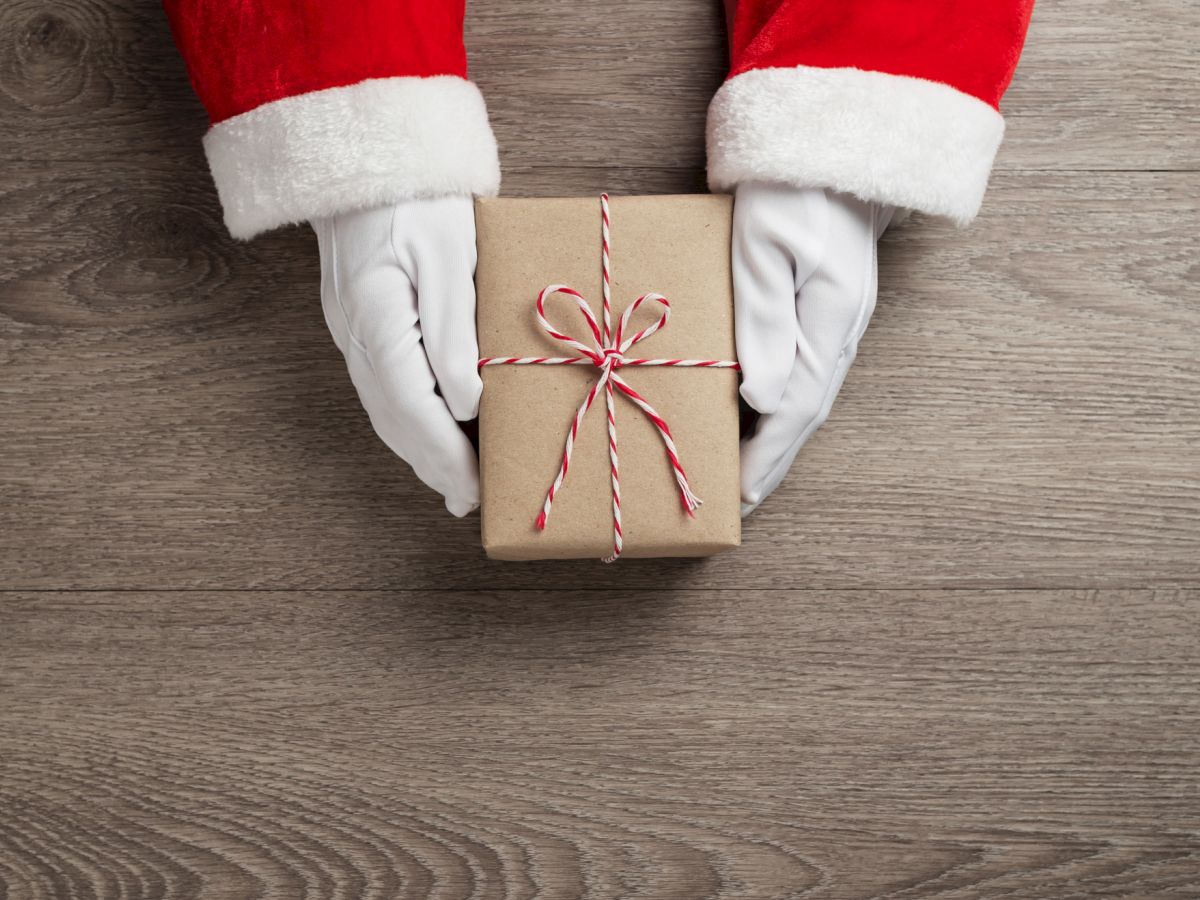 Santa Claus hands hold a small, gift-wrapped box tied with red and white string on a wooden surface.