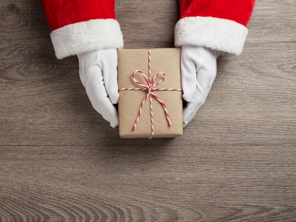 Santa-themed hands hold a small, brown gift box tied with red and white string on a wooden surface.