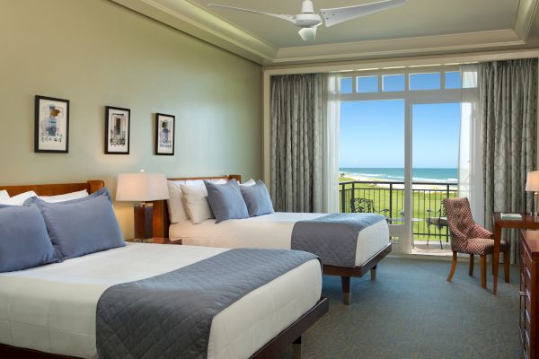 A hotel room with two beds, artwork on the wall, a desk with a chair, and a large window with an ocean view in the background.