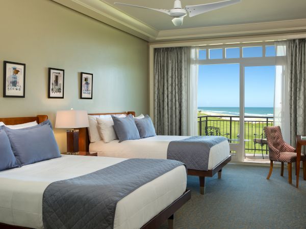 A hotel room with two beds, artwork on the wall, a desk with a chair, and a large window with an ocean view in the background.