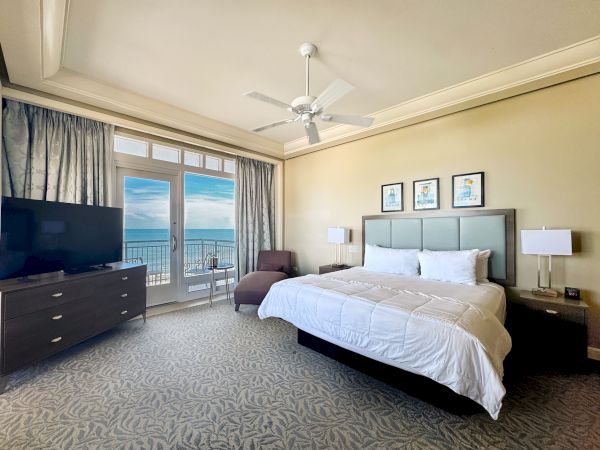 A modern hotel bedroom with a king bed, TV, lamps, ceiling fan, and a balcony view of the ocean and sky.