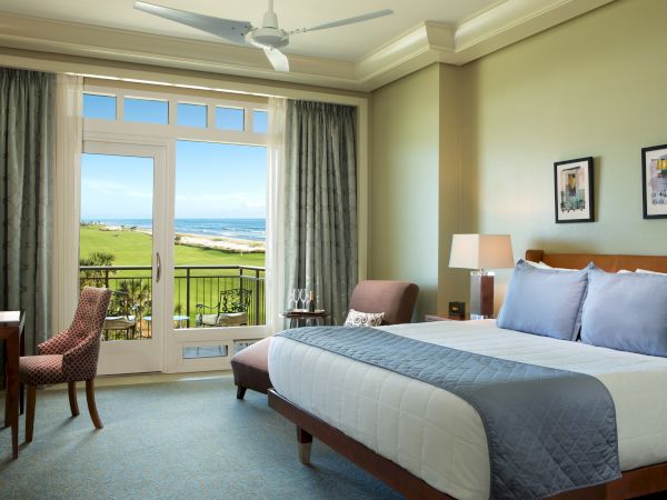 A hotel room with a king-sized bed, two lamps, a chair, and a desk; balcony view of the ocean and green golf course outside.