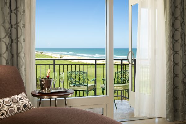 A cozy room with a view of a balcony overlooking a golf course and ocean, with champagne on a table and stylish chairs outside.