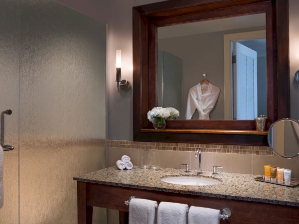 A modern bathroom vanity with a large mirror, neatly folded towels, floral decoration, toiletries, and a bathrobe reflected in the mirror.