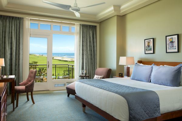 A cozy hotel room with a king-sized bed, balcony view of the ocean, desk, chair, and modern decor.